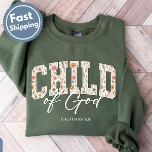 May include: A military green sweatshirt with the words "CHILD of God" in a floral design. The text "GALATIANS 3:26" is below the main text. A "Fast Shipping" sticker is in the upper left corner.