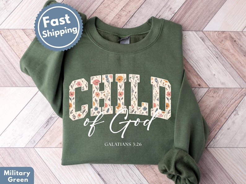 May include: A military green sweatshirt with the words "CHILD of God" in a floral design. The text "GALATIANS 3:26" is below the main text. A "Fast Shipping" sticker is in the upper left corner.