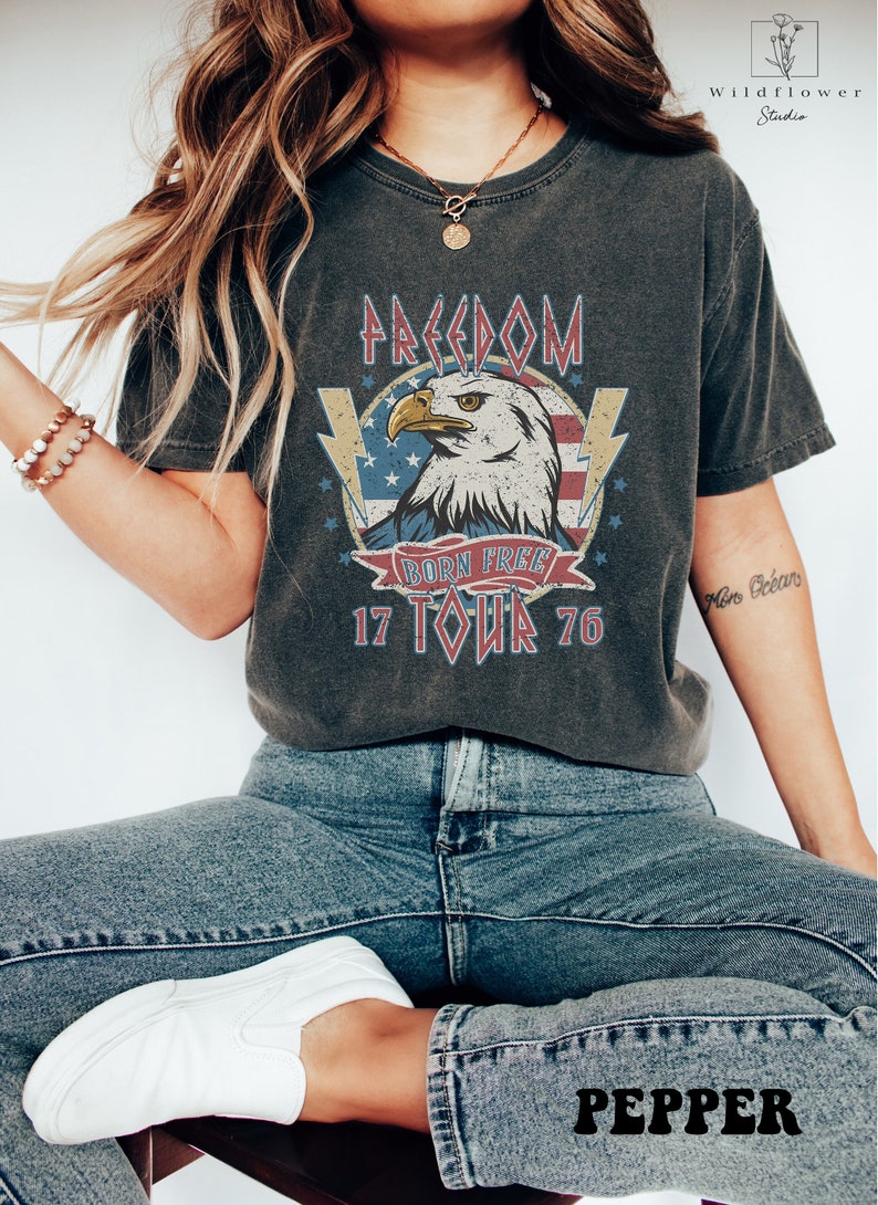 Puede incluir: Una camiseta gris oscuro con un gr&aacute;fico vintage de un &aacute;guila con las palabras "Freedom Born Free 17 Tour 76" impresas en ella.