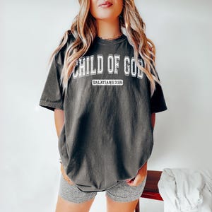 May include: A dark gray oversized t-shirt with the text "CHILD OF GOD" in white with a varsity-style font. The text "GALATIANS 3:26" is printed in white below the main text.