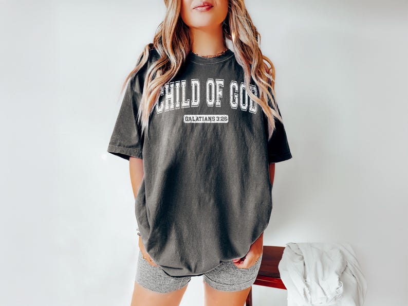 May include: A dark gray oversized t-shirt with the text "CHILD OF GOD" in white with a varsity-style font. The text "GALATIANS 3:26" is printed in white below the main text.