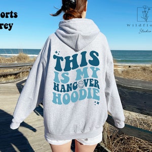May include: A gray hooded sweatshirt with the text "This is my hangover hoodie" in blue and teal lettering. The text is in a retro font with a smiley face graphic.