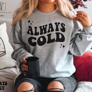 May include: A gray sweatshirt with the text "ALWAYS COLD" in a retro font. The text is in black and there are small stars on either side of the text.