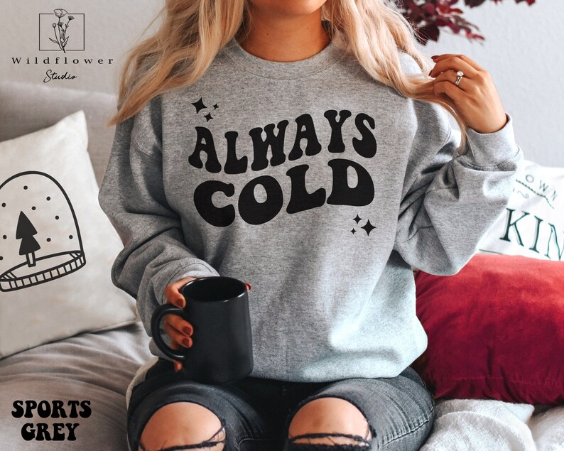 May include: A gray sweatshirt with the text "ALWAYS COLD" in a retro font. The text is in black and there are small stars on either side of the text.