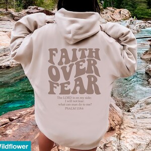 K&ouml;nnte beinhalten: Beigefarbener Kapuzenpullover mit dem Schriftzug "FAITH OVER FEAR" in braunen Retro-Lettern. Darunter steht der Bibelvers Psalm 118,6. Der Hoodie hat eine neutrale Farbe und ist vielseitig einsetzbar.