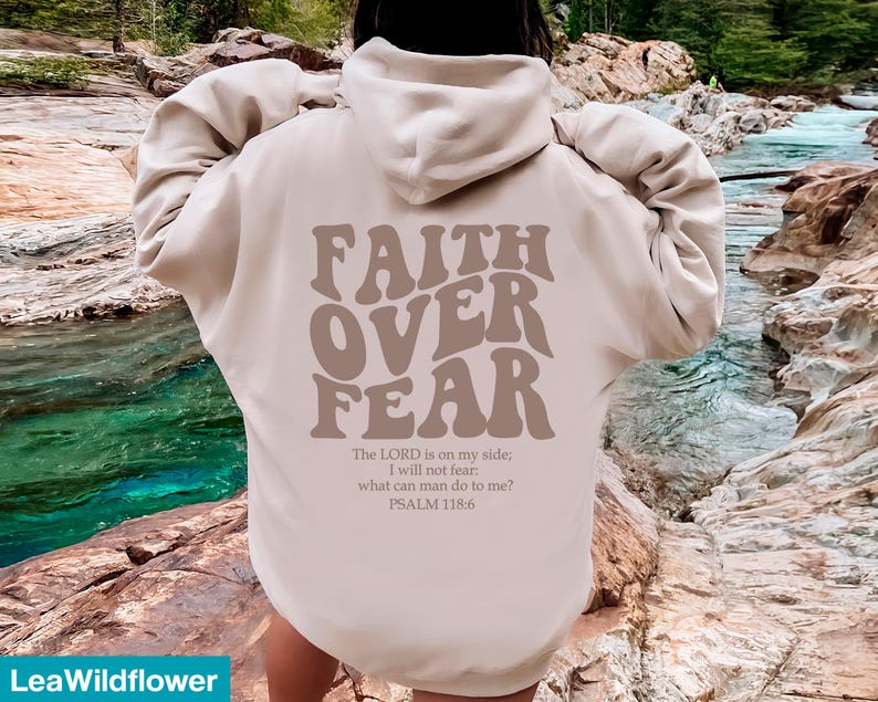K&ouml;nnte beinhalten: Beigefarbener Kapuzenpullover mit dem Schriftzug "FAITH OVER FEAR" in braunen Retro-Lettern. Darunter steht der Bibelvers Psalm 118,6. Der Hoodie hat eine neutrale Farbe und ist vielseitig einsetzbar.