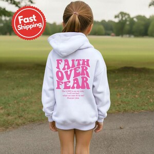 K&ouml;nnte beinhalten: Wei&szlig;er Kapuzenpullover mit dem pinkfarbenen Text "FAITH OVER FEAR" und einem Bibelvers. Der Hoodie hat eine Kapuze mit Kordelzug und lange &Auml;rmel. Der Text "Fast Shipping" befindet sich in einem roten Kreis oben links.