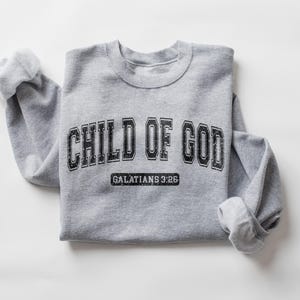 May include: Gray sweatshirt with the text "CHILD OF GOD" in black with a vintage collegiate font. Below the text is "GALATIANS 3:26" in a smaller font.