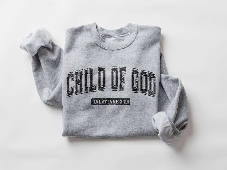 May include: Gray sweatshirt with the text "CHILD OF GOD" in black with a vintage collegiate font. Below the text is "GALATIANS 3:26" in a smaller font.