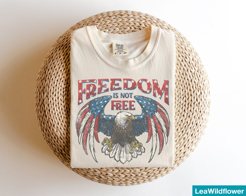 May include: A cream-colored t-shirt with a vintage-style graphic of an eagle with outstretched wings. The eagle is holding a banner that reads "Freedom is not free". The eagle is superimposed on a red, white, and blue American flag.