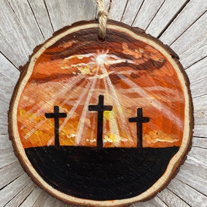May include: A wooden round ornament with a painted sunset scene and three black crosses silhouetted against the orange and yellow sky.