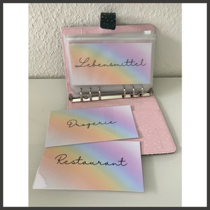 Puede incluir: Una carpeta rosa con un bolsillo de plástico transparente que contiene tres tarjetas de colores arcoíris. Las tarjetas tienen las palabras "Lebensmittel", "Drogerie" y "Restaurant" escritas en negro.