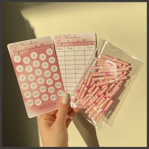 Op de afbeelding: Twee roze kaarten met de tekst "30 Wochen Sparlovechallenge" en een raster met de nummers 1-30. De kaarten liggen naast een doorzichtige plastic zak gevuld met roze en witte opgerolde papieren met roze en witte ontwerpen.