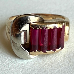 May include: A gold and silver ring featuring a buckle-style design. The center of the ring displays four vertical, rectangular, deep red stones set within a gold frame, connected to a polished silver-toned metal loop that resembles a belt buckle.