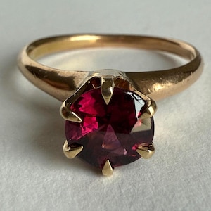 May include: A gold ring featuring a round, faceted, deep red gemstone set in a six-prong mounting. The band is smooth and polished, with a warm yellow gold tone that contrasts with the dark, saturated color of the central stone.