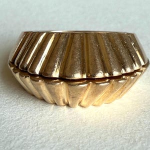 May include: A gold-colored, small, lidded trinket box with a fluted, Art Deco design. The box has a geometric shape with vertical ridges and a hinged lid. The box is made of a shiny, metallic material.