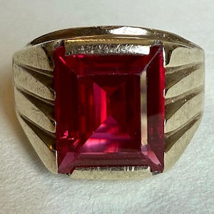 May include: A gold-toned ring featuring a large, rectangular, vibrant red gemstone. The ring's band has a ridged design on the sides, adding texture to the piece. The gemstone is the focal point, catching the light and showcasing its rich color.