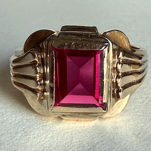 May include: A gold-toned ring featuring a large, rectangular, vibrant pink gemstone. The ring has a detailed design with ribbed accents on the sides and a decorative top. The gemstone is the focal point, set in a polished gold setting.