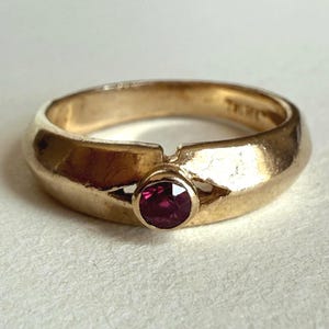 Peut inclure: Une bague en or avec une pierre précieuse rouge foncé et ronde au centre. La bague a un anneau épais et une finition lisse et polie. La pierre précieuse est d'une couleur vive et attrayante.