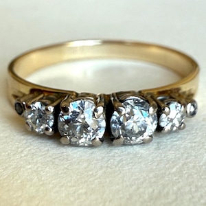 May include: A gold ring with four clear, round-cut gemstones set in a row. The ring's band is a simple, polished gold, and the gemstones are held in place by small prongs. The ring is likely a piece of jewelry, possibly an engagement or anniversary ring.