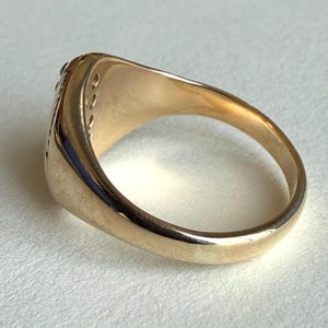 May include: A gold-colored ring with a rounded band and a flat top. The ring has a decorative design on the side and small, embedded stones along the top edge. The ring is shown on a light-colored surface.