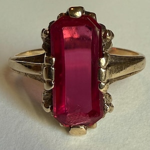 May include: A gold ring featuring a large, rectangular, faceted, deep pink gemstone set vertically. The metal band has a textured, organic-looking setting that secures the stone with multiple prongs, creating a bold and prominent centerpiece.