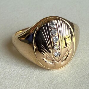 May include: A gold ring with a rounded face, featuring five small diamonds set vertically. The ring's face is engraved with a stylized design of radiating lines and floral elements.