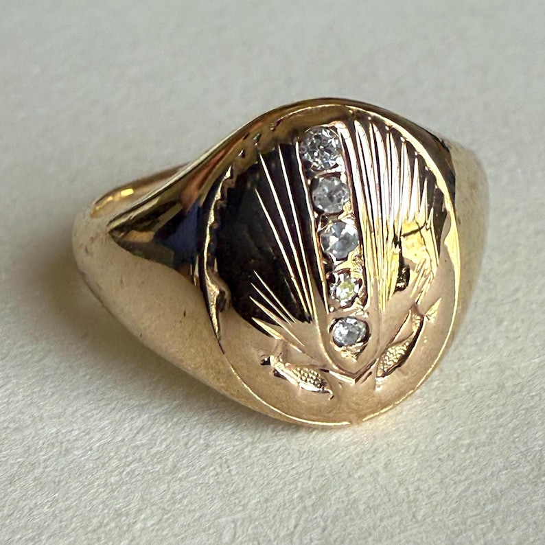 May include: A gold ring with a rounded face, featuring five small diamonds set vertically. The ring's face is engraved with a stylized design of radiating lines and floral elements.