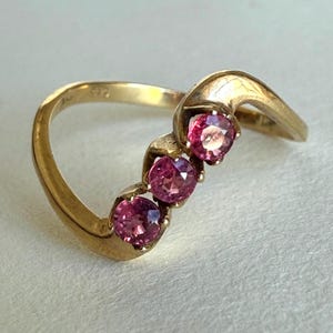 May include: A gold ring featuring three round, pink gemstones. The ring has a curved design with the gemstones set in a row. The gold band has a smooth, polished finish, and the gemstones are a vibrant pink color.