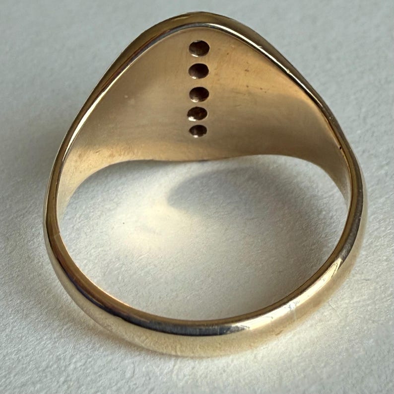 May include: A gold-colored ring with a rounded band and a flat, oval top. The top of the ring features six small, evenly spaced, dark-colored stones. The ring is set against a light-colored background.