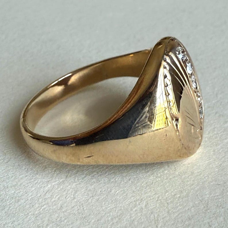 May include: A gold ring with a rounded, oval face. The face is adorned with small, clear gemstones and etched with a leaf-like design. The band is smooth and polished, reflecting light. A classic piece of jewelry.