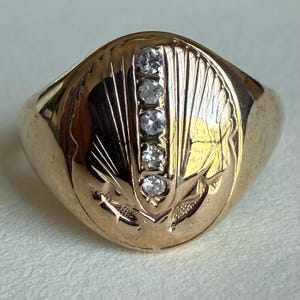 May include: A gold ring with a rounded face, featuring five small, clear gemstones set vertically. The ring has an engraved design of a stylized leaf or feather pattern on the face. The ring is a warm, golden color.