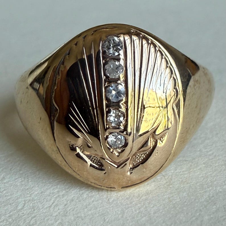 May include: A gold ring with a rounded face, featuring five small, clear gemstones set vertically. The ring has an engraved design of a stylized leaf or feather pattern on the face. The ring is a warm, golden color.