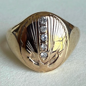 May include: A gold-colored ring with five clear, round gemstones set in a row. The ring has an engraved design on the face, featuring a stylized pattern. The band is smooth and polished.