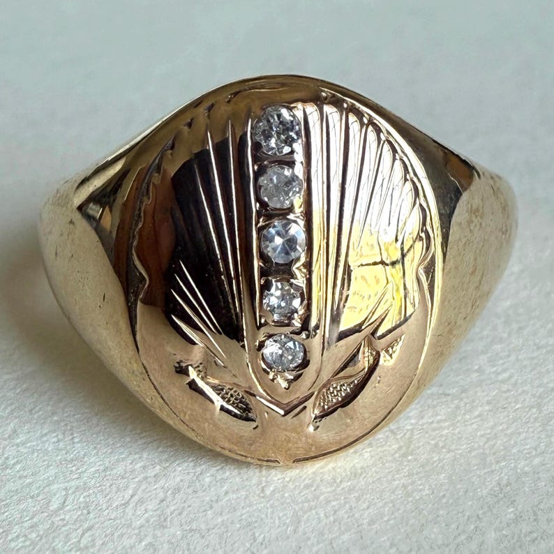 May include: A gold-colored ring with five clear, round gemstones set in a row. The ring has an engraved design on the face, featuring a stylized pattern. The band is smooth and polished.