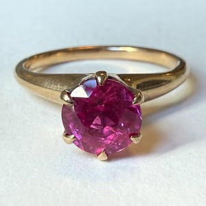 May include: A gold ring featuring a round, faceted, vibrant magenta-pink gemstone set in a six-prong mounting. The band is smooth and polished, tapering slightly toward the setting, which holds the circular stone securely in place.
