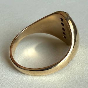 May include: A gold ring with a rectangular face, featuring a row of small, dark stones. The ring has a smooth, polished band and a slightly curved design. The ring is shown on a white surface.