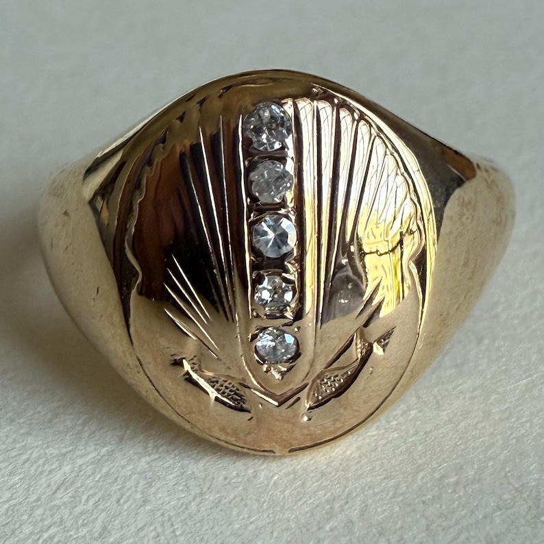May include: A gold-colored ring with a rounded band and a detailed oval face. The face features five clear, round gemstones set vertically, with etched details around them. The ring has an engraved design on the face.