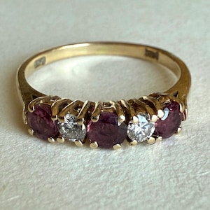 May include: A gold ring featuring five gemstones. The ring alternates between round, deep red gemstones and clear, round gemstones. The ring is set in a gold band.