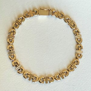 May include: A gold-toned bracelet with a repeating geometric design. The links are shaped like a stylized letter "G" connected by horizontal bars. The clasp is a simple rectangular design.