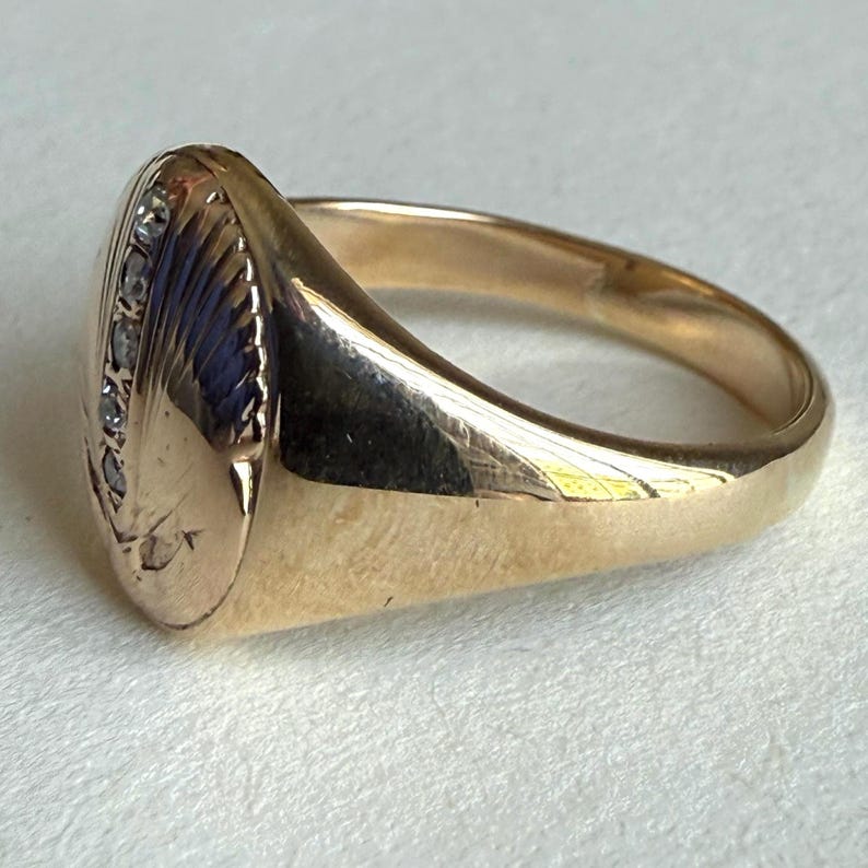 May include: A gold ring with a rounded, oval face. The face features a textured design with a row of small, clear stones along one edge. The band is smooth and polished, reflecting light.