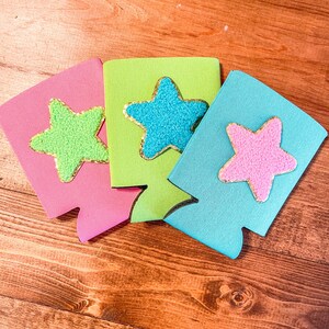 May include: Three colorful can coolers with star-shaped designs. The stars are made of a fuzzy material and have gold glitter accents. The coolers are pink, green, and blue.