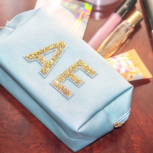 Peut inclure: Une trousse de maquillage zippée bleu clair avec les initiales "AE" en strass dorés.