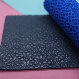 May include: A bright blue cylindrical rolling pin with a raised floral pattern and a flat, dark navy blue rectangular sheet of material featuring the same embossed flower design, showing the texture created by the roller on the surface.