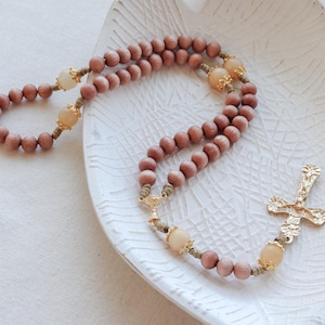 Puede incluir: Un rosario de madera con cuentas beige y una cruz dorada. El rosario está colocado sobre un plato de cerámica blanca con una superficie texturizada.