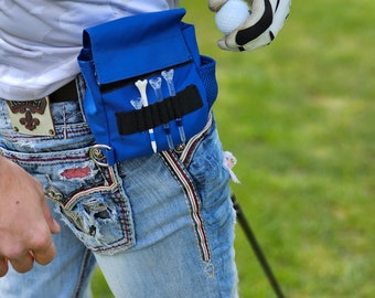 May include: A blue golf accessory pouch attached to a person's waist, containing golf tees. A golf ball is held in a white golf glove. The person is wearing light blue jeans. The background is a green grassy area.