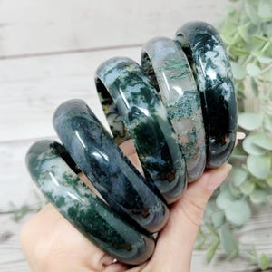 May include: A stack of five polished moss agate bangle bracelets featuring translucent and opaque sections with deep green, forest green, and white inclusions. Each circular stone bracelet displays unique natural patterns and a smooth, glossy finish.