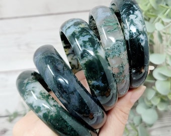 May include: A stack of five polished moss agate bangle bracelets featuring translucent and opaque sections with deep green, forest green, and white inclusions. Each circular stone bracelet displays unique natural patterns and a smooth, glossy finish.