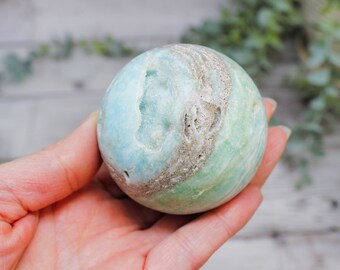 May include: A light blue and green sphere with a swirling pattern and white accents. The sphere is made of a natural material, possibly a type of agate or jasper.