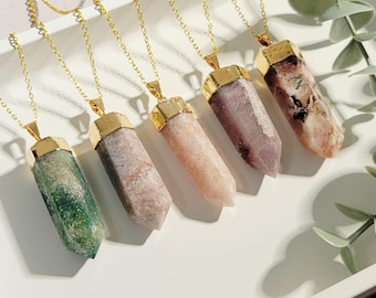May include: Five gold-toned necklaces with crystal pendants in shades of green, pink, and purple. Each pendant has a gold cap and hangs from a delicate gold chain. The necklaces are displayed on a white tray with greenery.